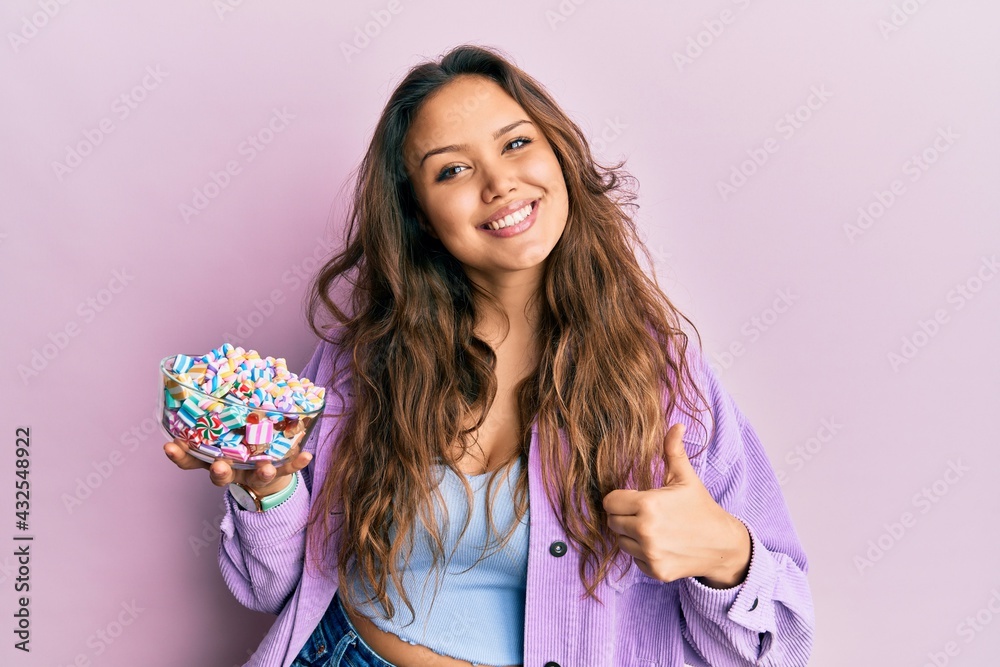 Wall mural young hispanic girl holding bowl of sugar candy smiling happy and positive, thumb up doing excellent