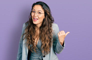 Young hispanic girl wearing business clothes and glasses smiling with happy face looking and pointing to the side with thumb up.