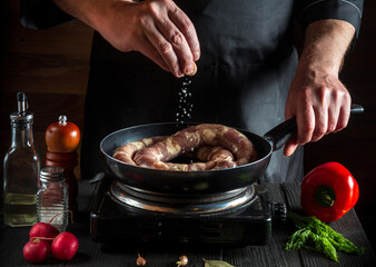 Professional chef or cook adds salt to pan with raw meat sausage. Preparation for cooking sausages in the kitchen of a restaurant or cafe on a table with vegetables and spices