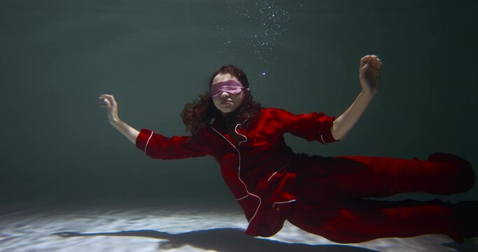 FALLING ASLEEP. Young beautiful woman wearing red pyjamas, sleep mask floats deep under water with open arms slow motion