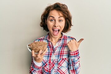 Young brunette woman holding bowl with lentils pointing thumb up to the side smiling happy with...
