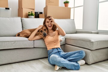 Obraz premium Young caucasian girl wearing casual clothes sitting on the floor with dog at home smiling happy doing ok sign with hand on eye looking through fingers