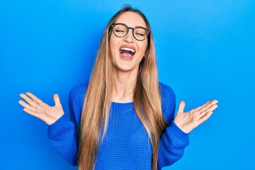 Fototapeta premium Young blonde girl wearing casual clothes and glasses celebrating mad and crazy for success with arms raised and closed eyes screaming excited. winner concept