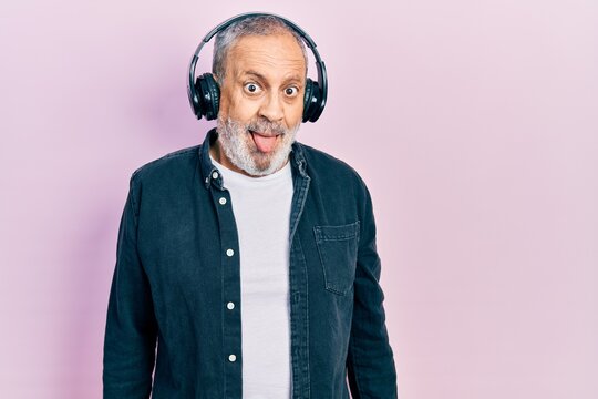 Handsome Senior Man With Beard Listening To Music Using Headphones Sticking Tongue Out Happy With Funny Expression. Emotion Concept.