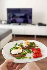 Young woman close up of her hand holding a plate with breakfast - pouched egg on whole grain toasted bread with cherry tomatoes and green pepper, she is sitting in living room watching TV, relaxing