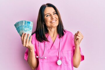 Middle age brunette doctor woman holding 100 brazilian reals banknotes screaming proud, celebrating victory and success very excited with raised arm