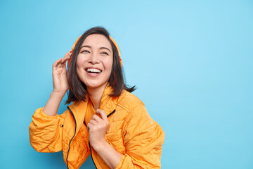Positive Asian woman enjoys favorite music in headphones smiles broadly expresses happiness wears...