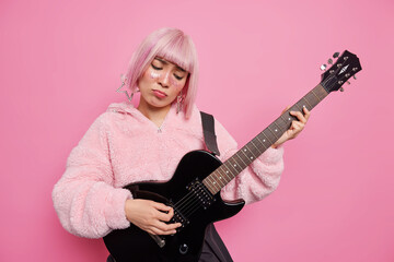 Obraz premium Displeased Asian woman plays solo on electric guitar wears warm coat has glitters on face pink bob hair looks sadly performs music isolated rosy background. Talented musician upset about something