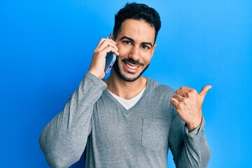 Young hispanic man having conversation talking on the smartphone pointing thumb up to the side smiling happy with open mouth