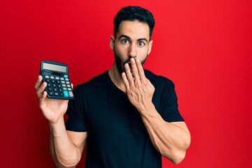 Young hispanic man showing calculator device covering mouth with hand, shocked and afraid for...