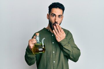 Young hispanic man holding olive oil can covering mouth with hand, shocked and afraid for mistake....