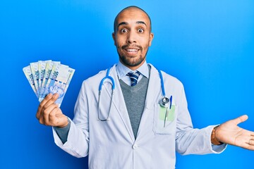 Hispanic adult doctor man wearing medical uniform holding 100 south african rand celebrating achievement with happy smile and winner expression with raised hand