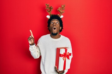 Young african american man wearing deer christmas hat holding gift amazed and surprised looking up and pointing with fingers and raised arms.