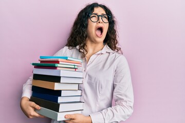Young brunette woman with curly hair holding a pile of books angry and mad screaming frustrated and furious, shouting with anger looking up.