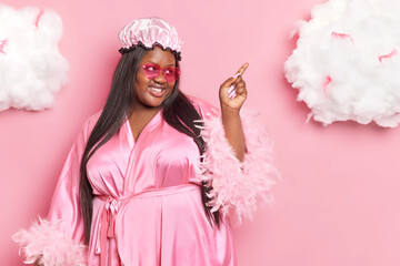 Horizontal shot of pleased dark skinned obese woman wears shower cap and dressing gown indicates at white cloud poses against pink background. Overweight dark skinned female wears home clothes
