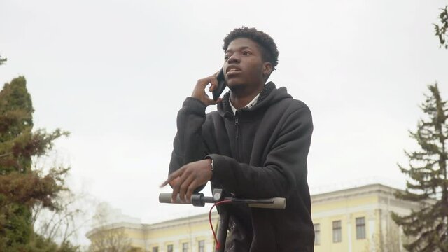 A young African-American man in a black sweater and black jeans and sneakers leans on an electric scooter and talks on a cell phone