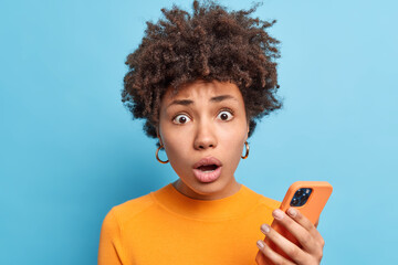 Concerned shocked woman with dark curly hair holds smartphone receives surprising news in message stands amazed and thrilled wears casual orange jumper isolated over blue background. Omg concept