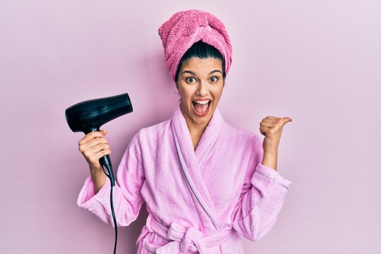Young hispanic woman wearing shower bathrobe using dryer pointing thumb up to the side smiling happy with open mouth