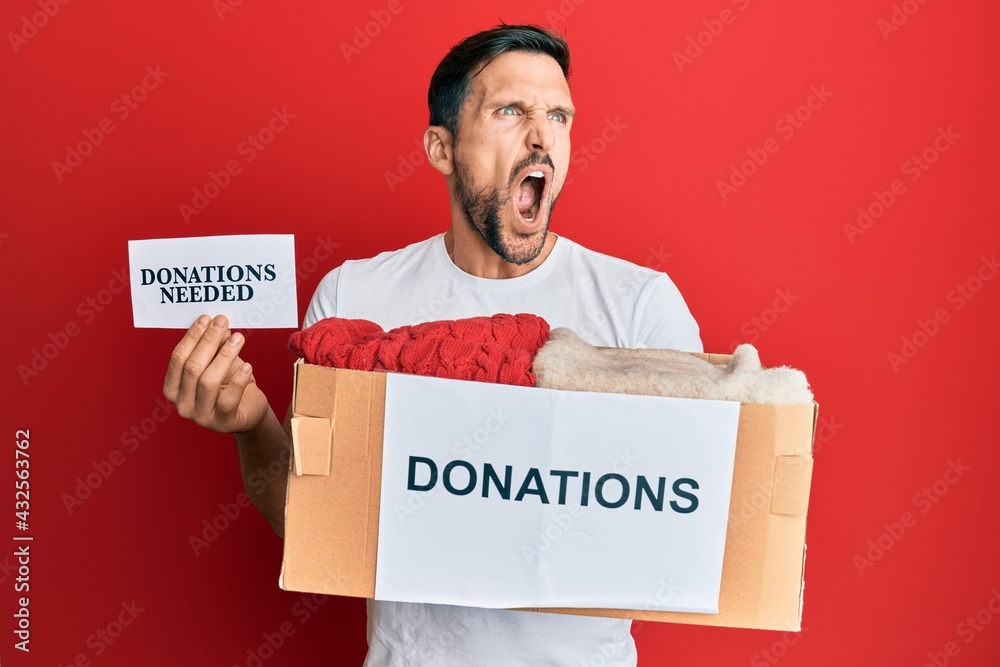 Poster Young handsome man volunteer holding box and donations needed paper angry and mad screaming frustrated and furious, shouting with anger. rage and aggressive concept.