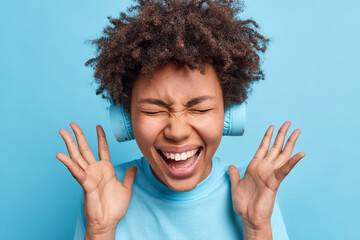 Overjoyed Afro American woman keeps hands raised exclaims joyfully closes eyes from happiness reacts on awesome news wears wireless headphones on ears isolated over blue background. Joy concept