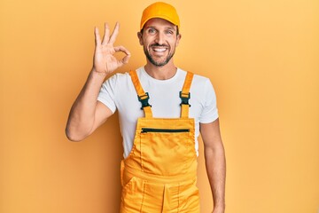 Young handsome man wearing handyman uniform over yellow background smiling positive doing ok sign with hand and fingers. successful expression.
