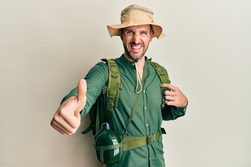 Fototapeta premium Handsome man with beard wearing explorer hat and backpack approving doing positive gesture with hand, thumbs up smiling and happy for success. winner gesture.