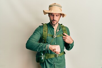 Fototapeta premium Handsome man with beard wearing explorer hat and backpack checking the time on wrist watch, relaxed and confident