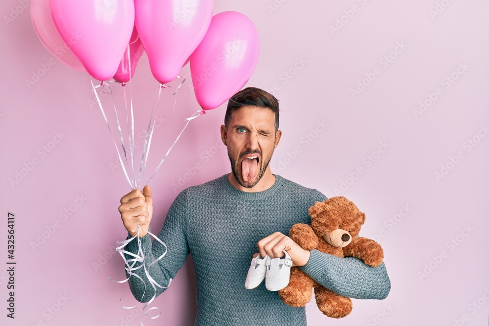 Sticker Handsome man with beard expecting a baby girl holding balloons, shoes and teddy bear sticking tongue out happy with funny expression.