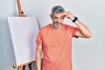 Handsome middle age man with grey hair standing by painter easel stand worried and stressed about a...