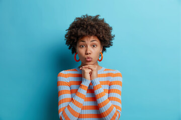 Fototapeta na wymiar Horizontal shot of lovely curly haired African American woman keeps hands under chin lips folded wears striped jumper wants to kiss you poses against blue background. Facial expressions concept
