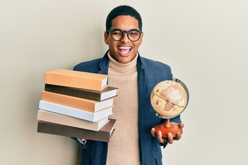 Young handsome hispanic man holding vintage world ball and pile of books winking looking at the...