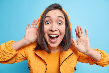 Excited suprised cheerful young Asian woman keeps mouth opened wears headphones on ears listens audio track being shocked and very happy dressed in orange jacket isolated over blue background