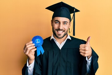 Young hispanic man wearing graduation robe holding 1 st place badge smiling happy and positive,...