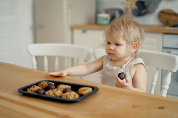 Little cute blonde chef girl playing in the kitchen. Eats sweet delicious candy and dessert. A healthy child and a cheerful child's mood. Bright kitchen in the apartment.