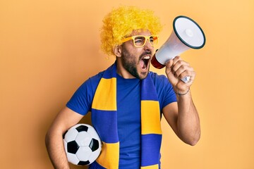 Young hispanic man football supporter holding soccer ball using megaphone angry and mad screaming frustrated and furious, shouting with anger. rage and aggressive concept.
