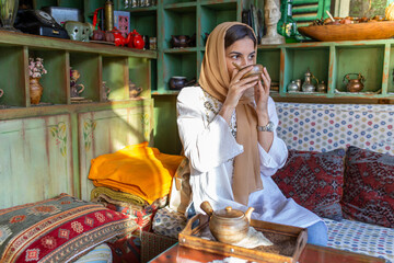 Young smiling muslim woman with hijab is sitting in an oriental tea bar and drinking a hot beverage.
