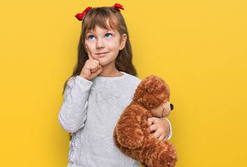 Little caucasian girl kid hugging teddy bear stuffed animal serious face thinking about question with hand on chin, thoughtful about confusing idea