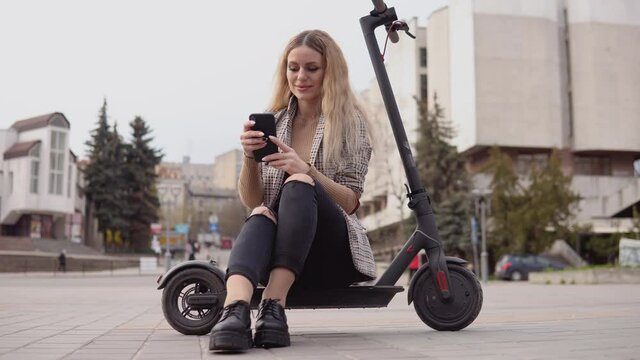 Young stylish blonde girl in a light jacket and jeans sits on an electric scooter and prints in a cell phone on the background of a quiet street