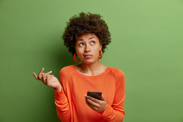 Perplexed hesitant curly haired African American woman shrugs shoulder being not sure uses smartphone device concentrated upwards wears casual orange jumper isolated over green studio background.