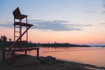 Fototapeta na wymiar Silhouette of an empty red lifeguard chair during idyllic sunset or sunrise at Lamure Beach along the Ottawa River at Deep River, Ontario Canada.