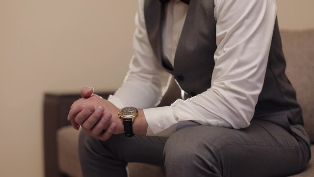 Young man in suit sitting on sofa with wrist watch clock on hand. Groom dresses. Handsome male businessman is waiting for a meeting. Wristwatch on bridegroom arm. Wedding morning. Slow motion. Backlit