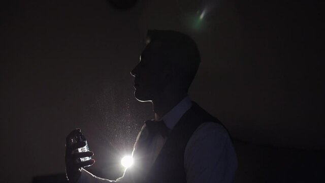 Groom splits the perfume or deodorant on hisself. Wedding day. Slow motion. Well dressed young adult handsome businessman using perfume from a bottle on his neck in dark room background. Backlit