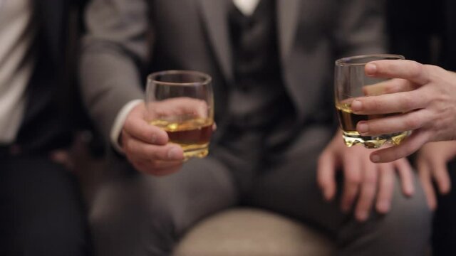Company of grooms friends sitting, talking, holding and clinking glasses of alcoholic whiskey drinks in their hands. Male hands are holding glasses of cognac, brandy, close-up. Celebrate wedding day