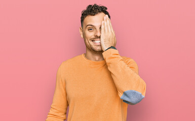 Young hispanic man wearing casual clothes covering one eye with hand, confident smile on face and surprise emotion.