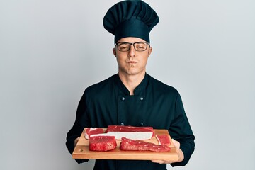 Handsome young man wearing chef uniform holding board with raw meat puffing cheeks with funny face. mouth inflated with air, catching air. - obrazy, fototapety, plakaty