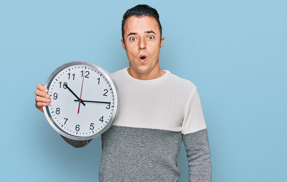 Handsome young man holding big clock scared and amazed with open mouth for surprise, disbelief face
