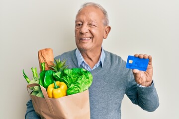 Senior caucasian man holding groceries and credit card winking looking at the camera with sexy...