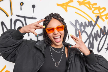 Cheerful cool ethnic woman with dreadlocks makes yo gesture has fun dressed in black jacket and...