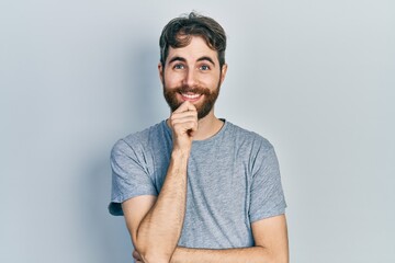 Caucasian man with beard wearing casual grey t shirt looking confident at the camera with smile with crossed arms and hand raised on chin. thinking positive.