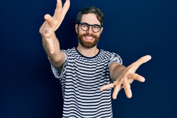 Naklejka premium Caucasian man with beard wearing striped t shirt and glasses looking at the camera smiling with open arms for hug. cheerful expression embracing happiness.